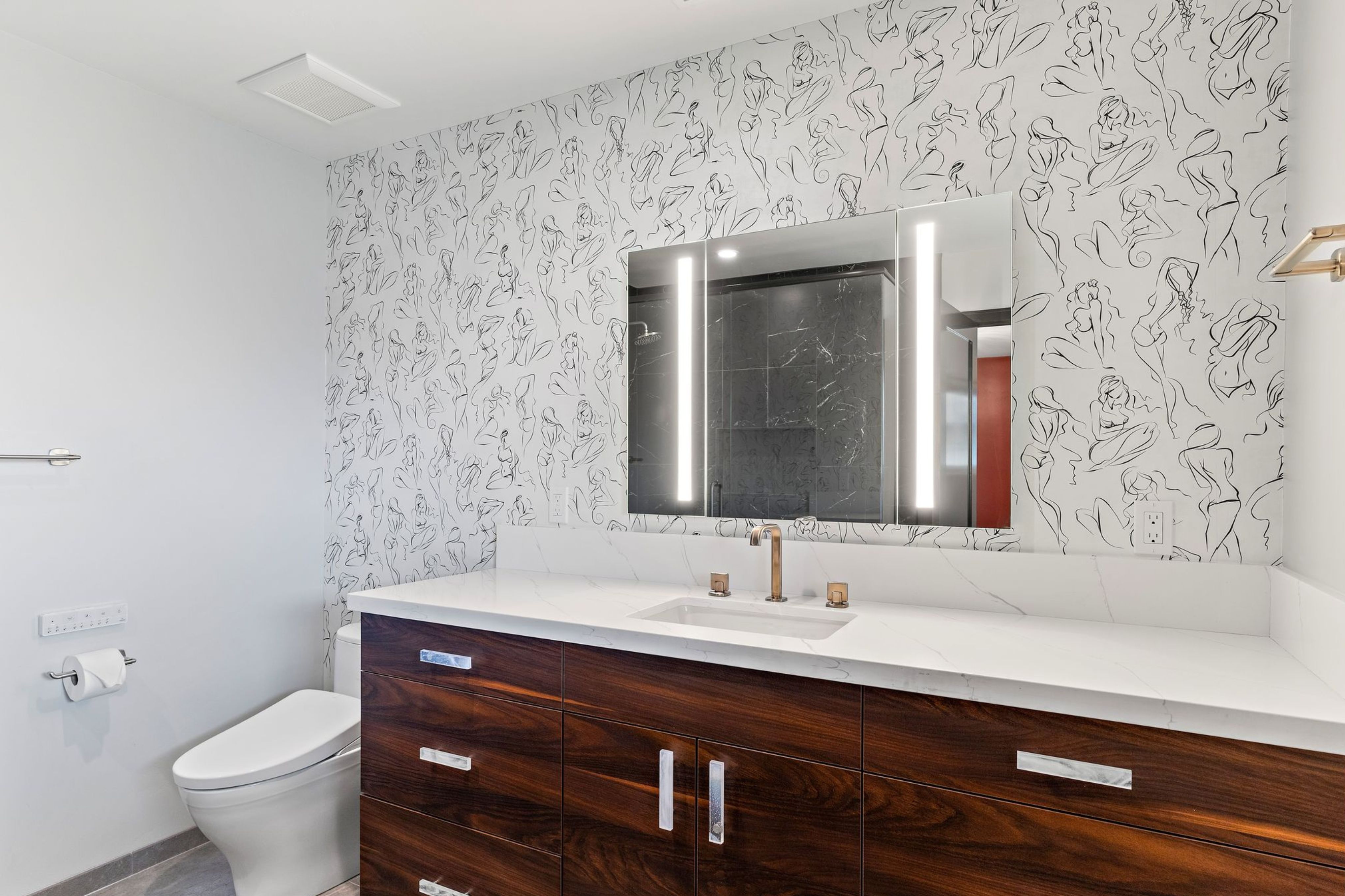 Primary bathroom with dark wood vanity, white countertop, illuminated mirror and line-art wallpaper