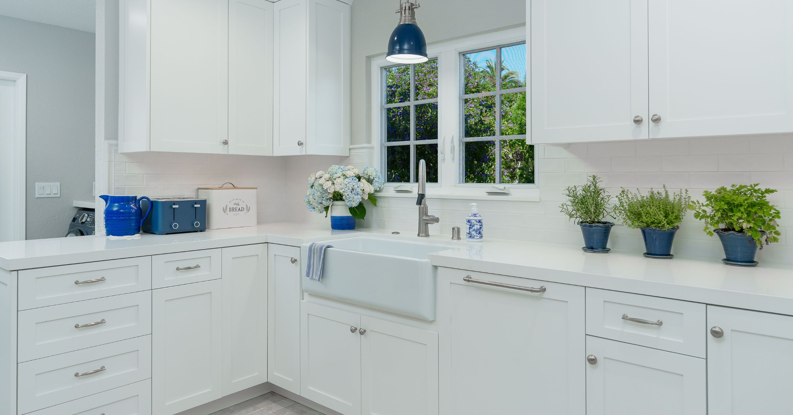 Farmhouse sink under two windows with white shaker cabinets, quartz counters, and blue pendant lighting.