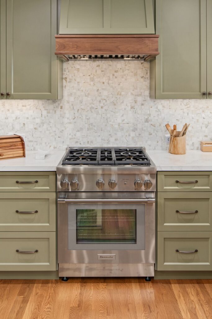 Stainless range beneath a sage and dark wood hood, surrounded by green cabinetry and mosaic tile backsplash.