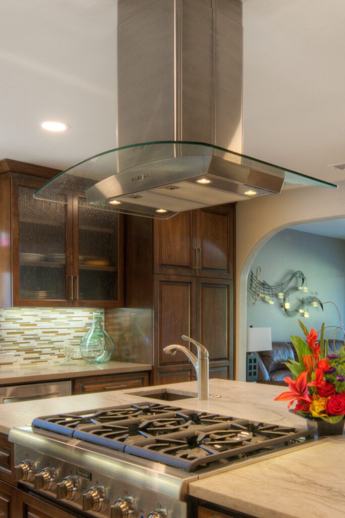 Curved glass and stainless steel island range hood above cooktop with dark wood cabinetry in the background.