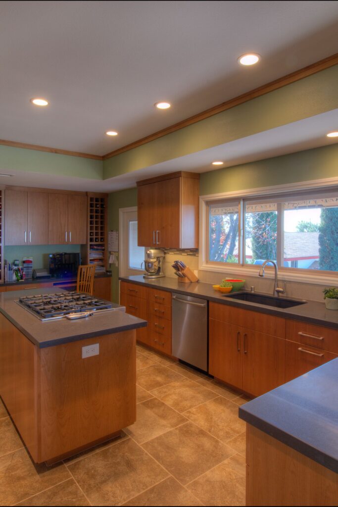 Kitchen with cooktop island, wood cabinets, stainless dishwasher, wide sink window and built-in desk area