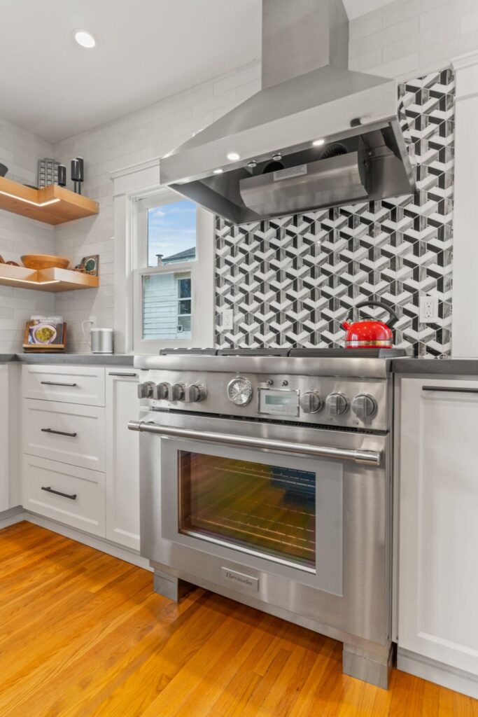 Kitchen cooking wall with stainless range and hood, geometric backsplash, floating wood shelves and white cabinets.