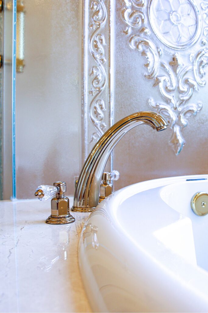Gold tub faucet with crystal handles beside a white soaking tub and decorative frosted glass panel.