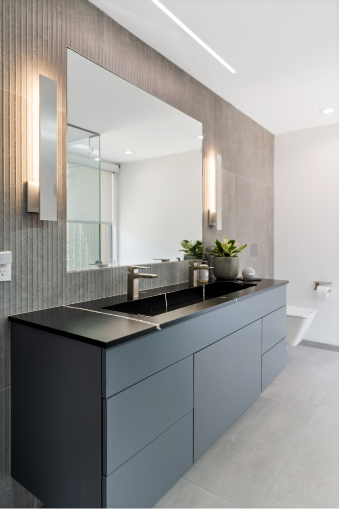 Bathroom vanity with gray flat-panel cabinetry, black countertop, large mirror, textured wall tile and vertical sconces.