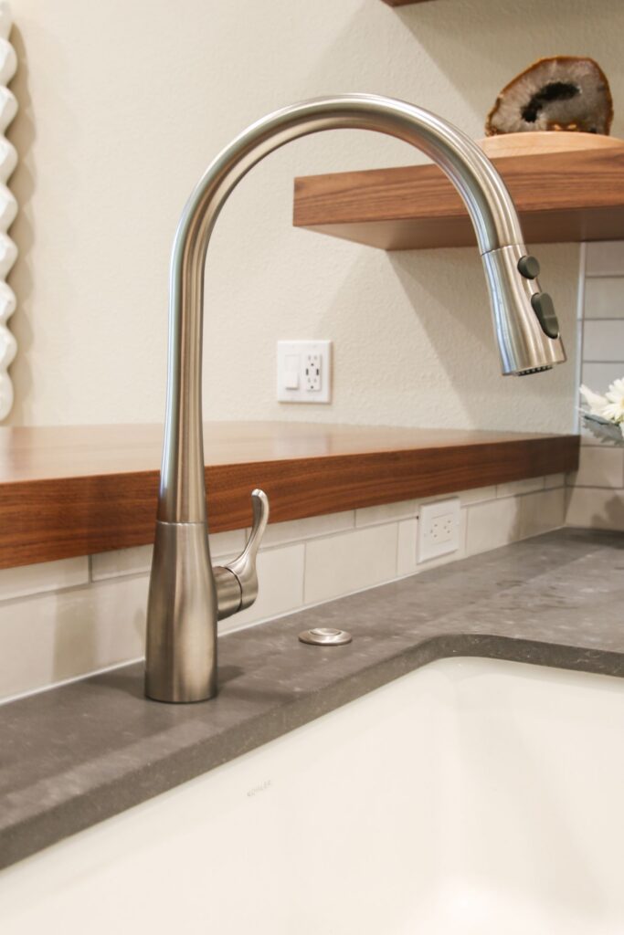 Pull-down stainless kitchen faucet over a white sink with gray countertop, wood ledge and floating shelf