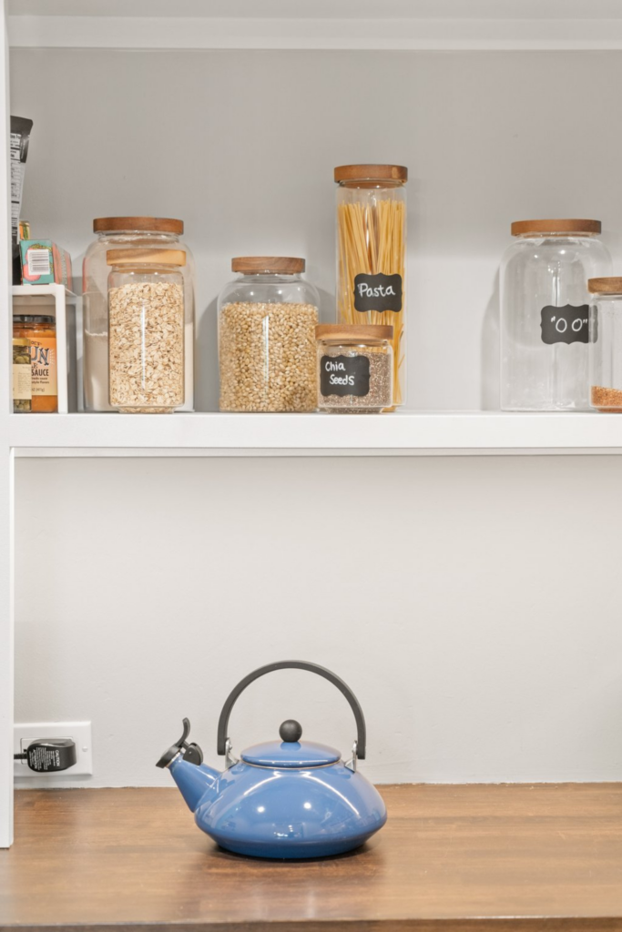 Pantry shelving with glass dry-goods jars above a wood countertop and blue kettle.