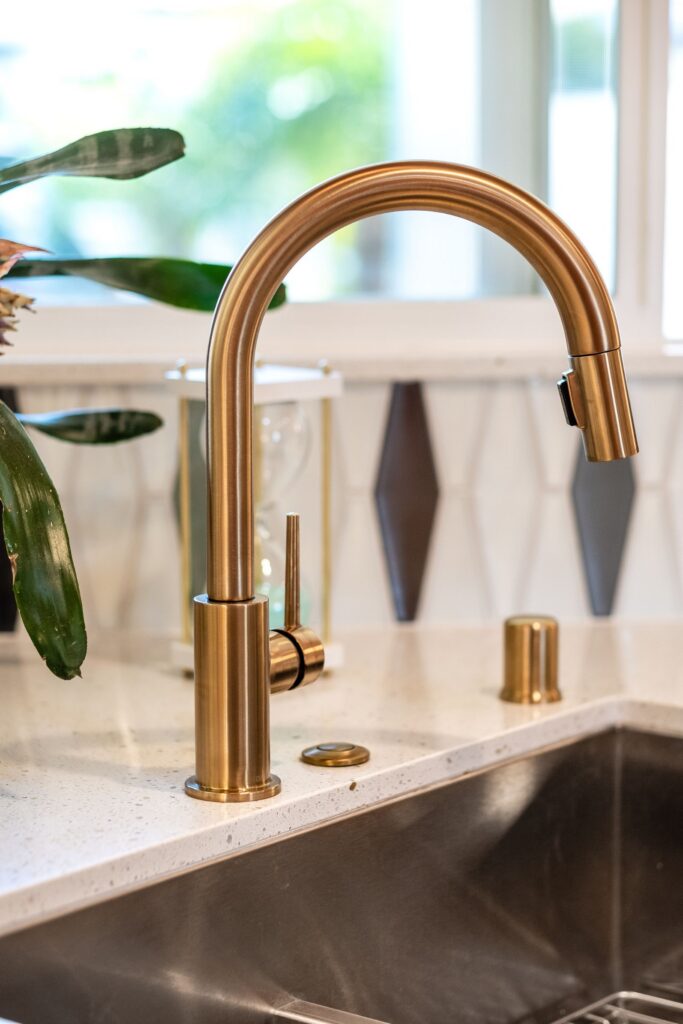 Brushed brass kitchen faucet over stainless sink with white countertop and geometric tile backsplash