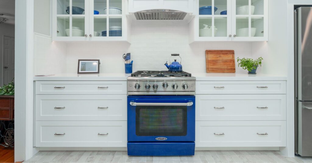 Centered blue gas range with white shaker drawers, glass cabinets, subway tile, and stainless hood insert.