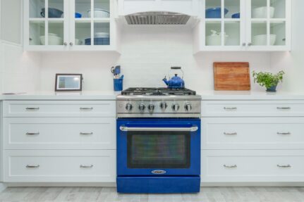 Centered blue gas range with white shaker drawers, glass cabinets, subway tile, and stainless hood insert.