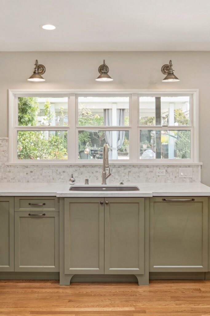 Kitchen sink wall with sage base cabinets, white counters, mosaic backsplash, wide windows, and three wall lights.