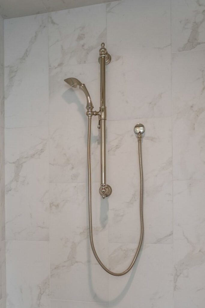 Bathroom shower wall with large marble-look tile and polished metal handheld shower fixture on an adjustable rail.