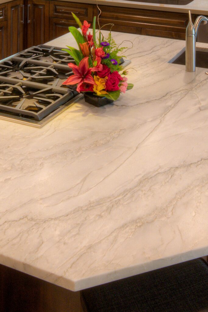 Light stone-look kitchen island countertop with integrated cooktop and prep sink beside dark wood cabinetry.