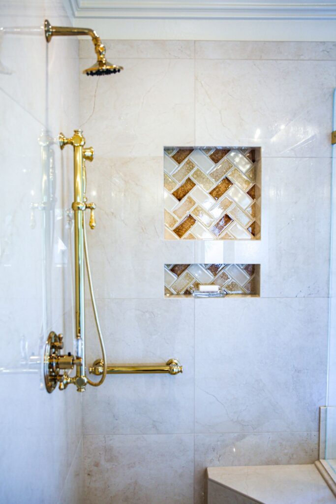 Gold shower fixtures with rain head, handheld spray and two mosaic-lined niches set in light stone tile.