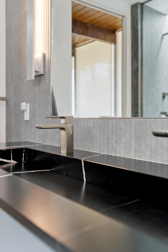 Bathroom sink detail with black stone countertop, brushed metal faucet, large mirror and textured gray backsplash.