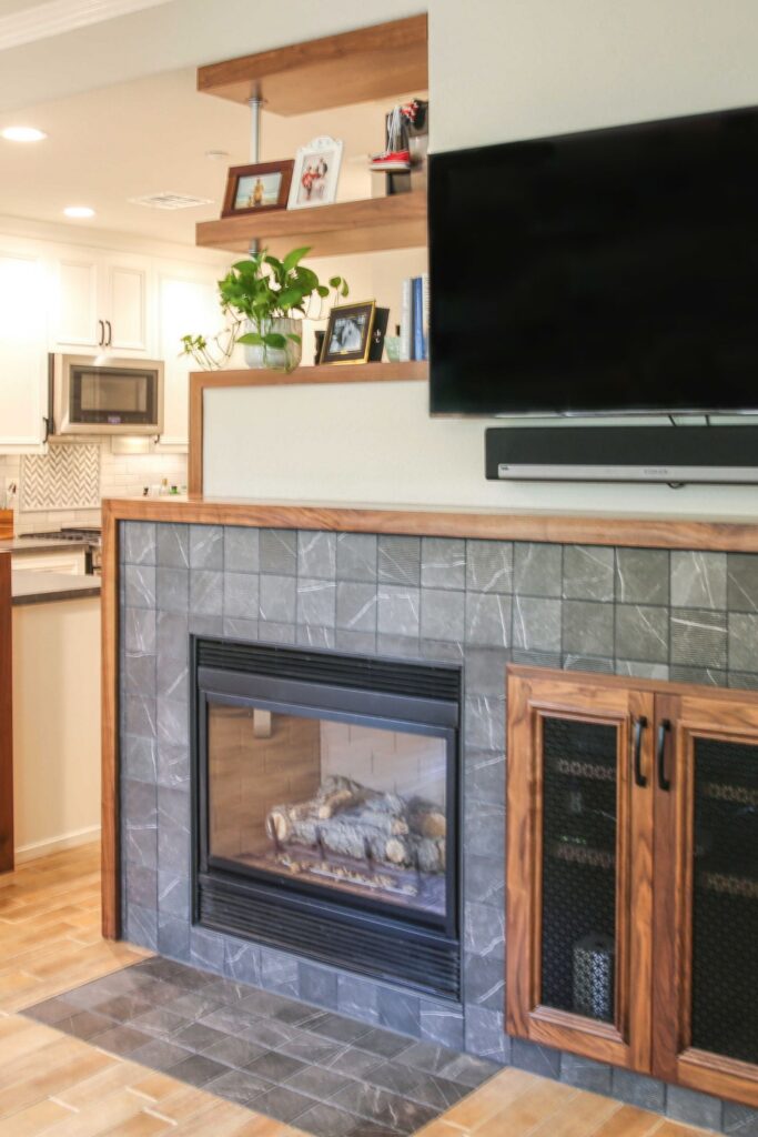 Living room fireplace with gray tile surround, wood trim, built-in cabinet, floating shelves and wall-mounted TV