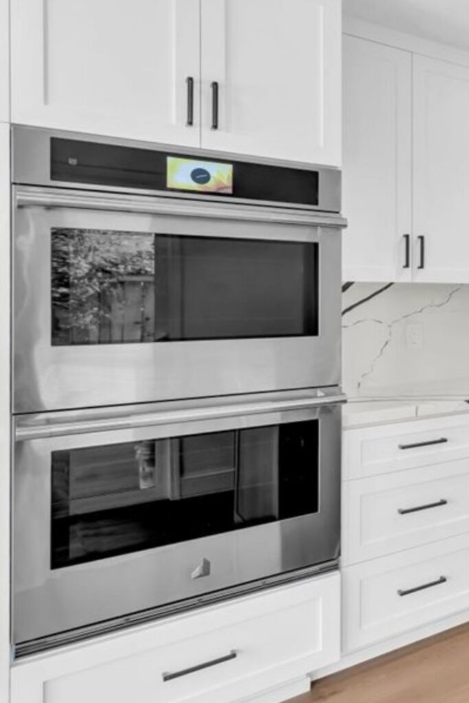 Stacked stainless wall ovens built into white cabinetry with black pulls and adjacent veined backsplash.