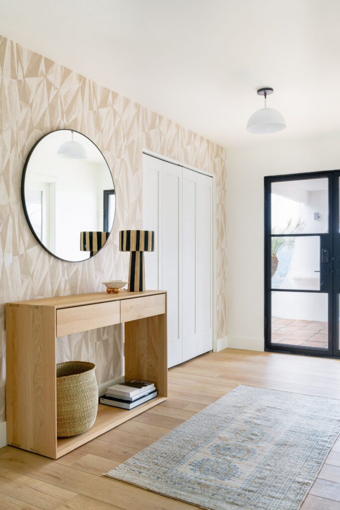 Entryway with patterned wallcovering, round black-framed mirror, light wood console and black-framed glass door