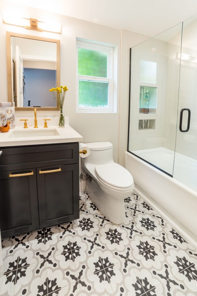 Bathroom with dark vanity, brass fixtures, patterned black-and-white floor tile and glass-enclosed tub shower