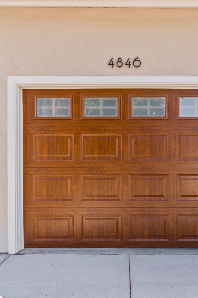 Wood-tone paneled garage door with upper windows, white trim and house numbers on stucco exterior