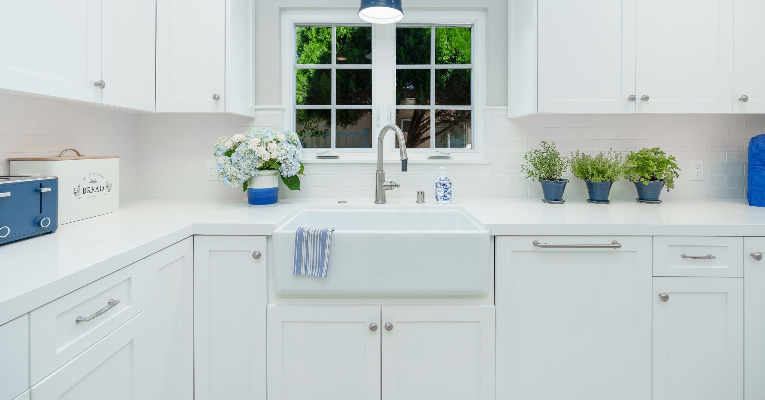 White farmhouse sink with pull-out faucet, shaker cabinetry, quartz counters, and garden-facing window.