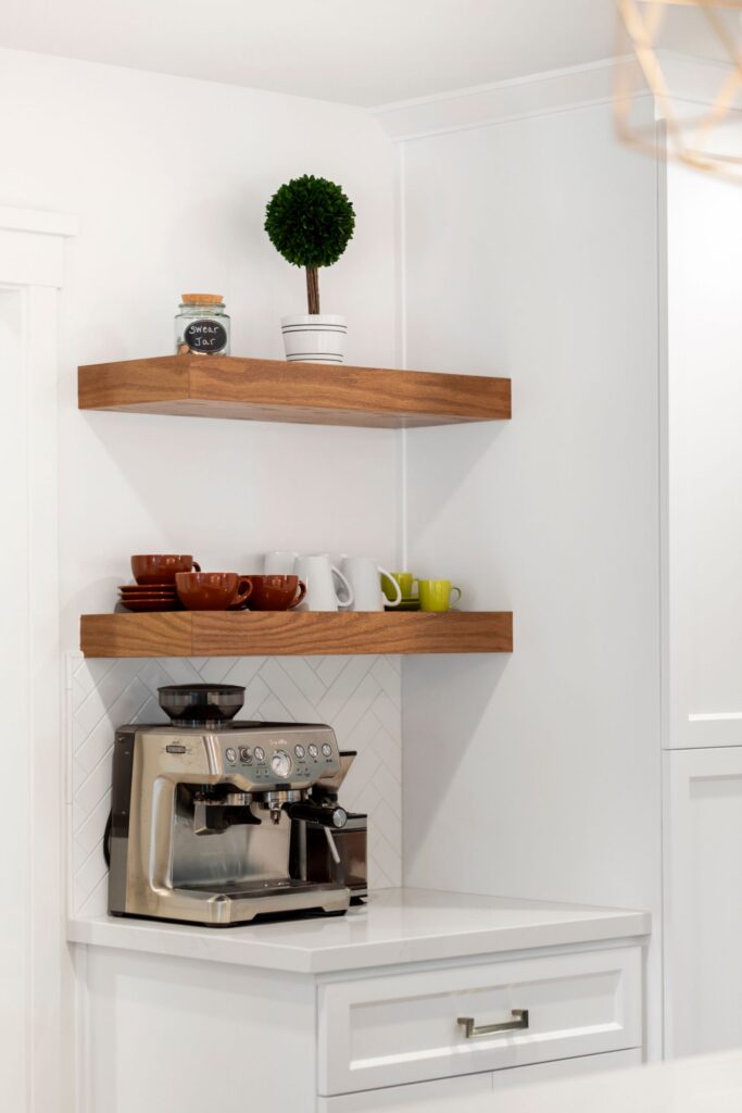 Coffee station with espresso machine, wood floating shelves, white cabinetry, and herringbone tile backsplash.