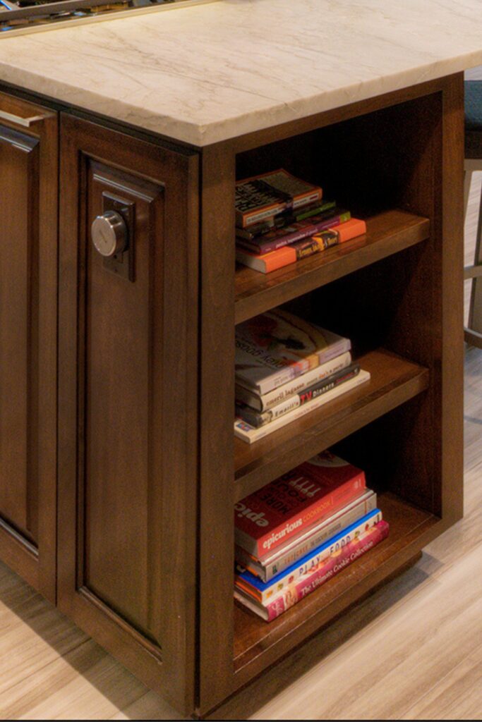 Open cookbook shelving built into the end of a dark wood kitchen island beneath a light stone-look countertop.