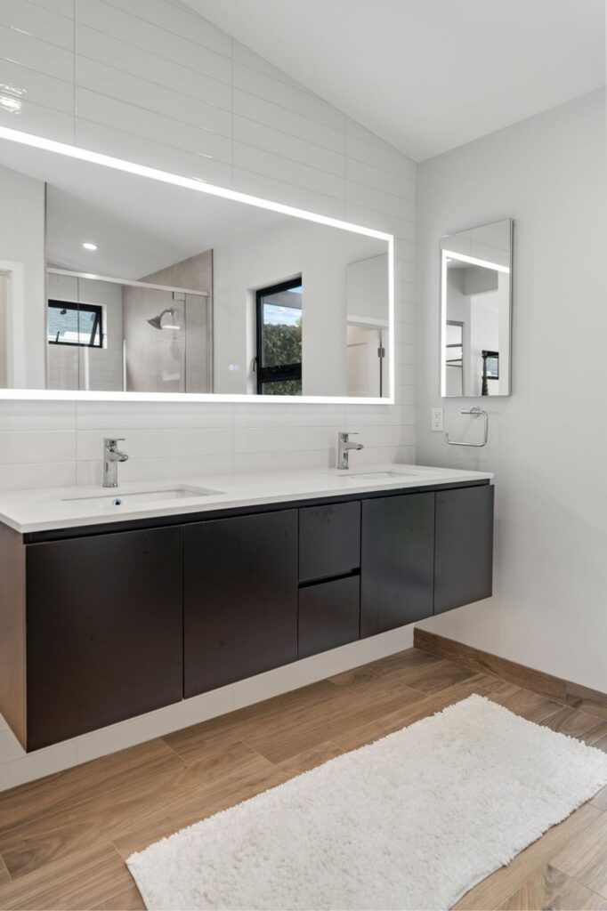 Double vanity with dark floating cabinetry, illuminated mirror, white horizontal wall tile and wood-look flooring.