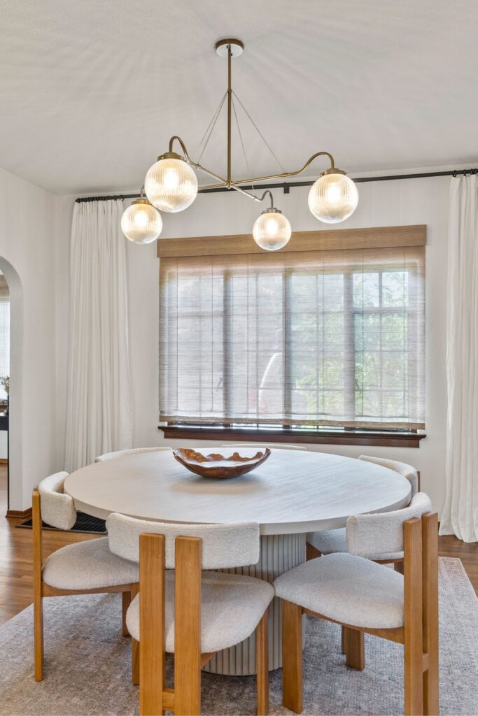 Dining room with round table, upholstered wood chairs, multi-globe chandelier, Roman shade and full-length curtains