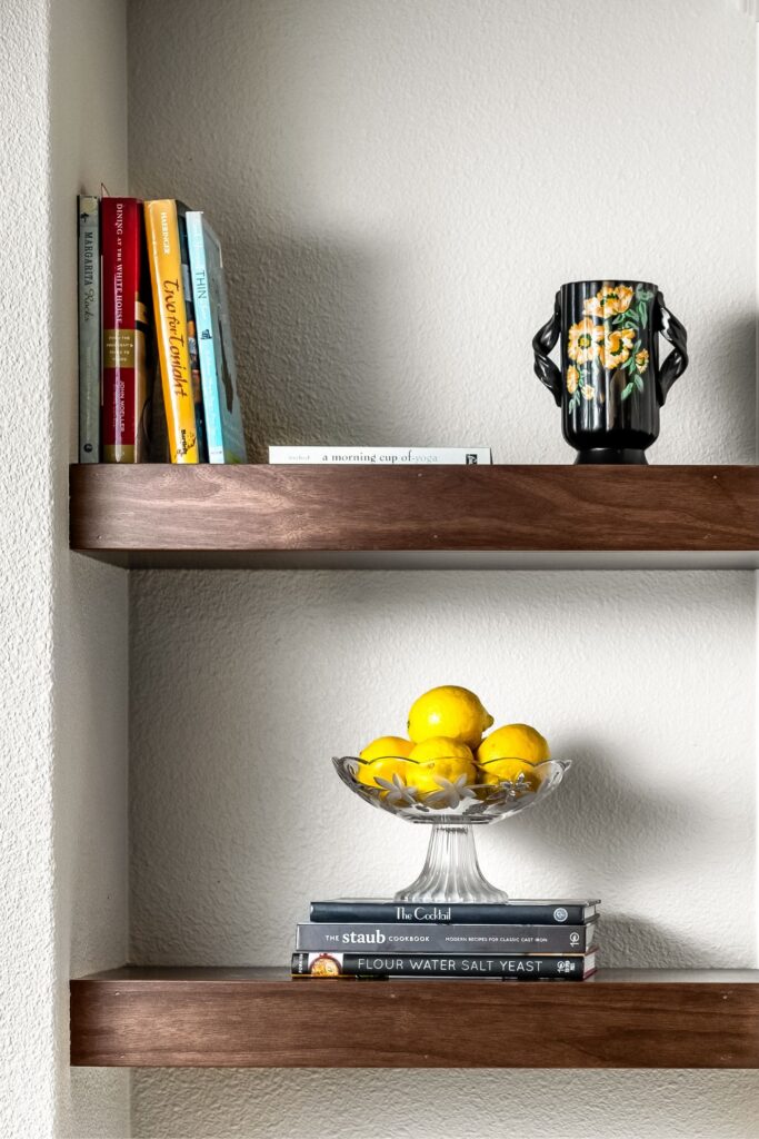 Floating dark wood kitchen shelves displaying cookbooks, decorative pottery and a glass bowl of lemons.