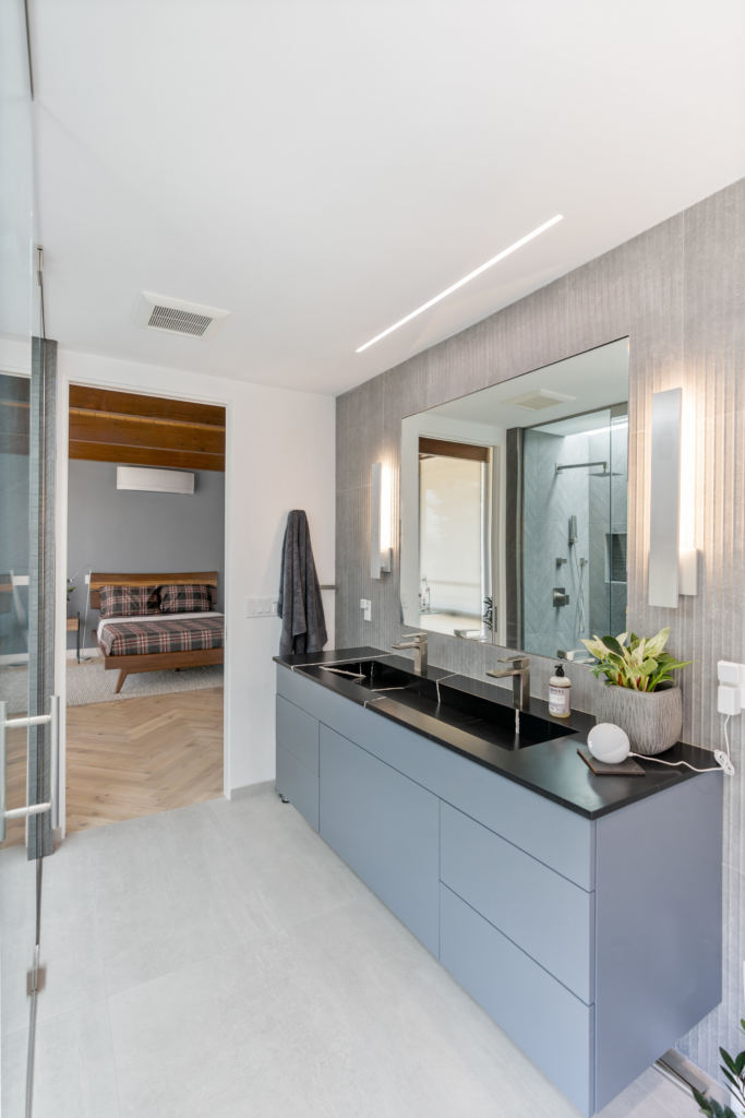 Primary bathroom vanity with gray cabinetry, black countertop, dual faucets, large mirror and vertical wall lighting.