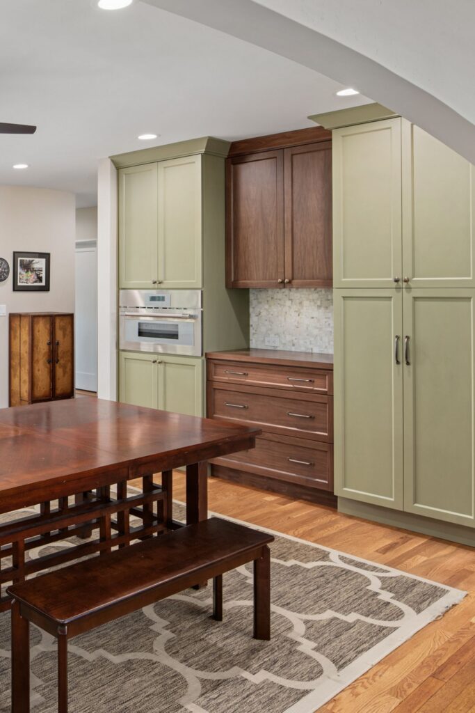 Dining area beside tall sage pantry cabinets, dark wood storage, wood table and bench, and an arched opening.