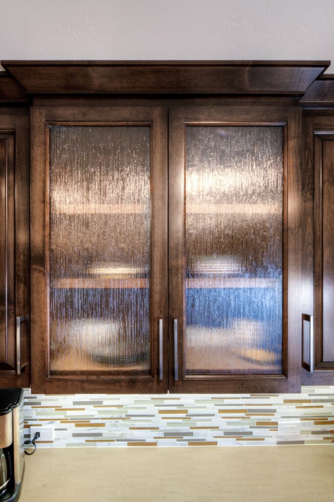 Textured glass upper cabinets framed in dark wood above a linear mosaic backsplash and light countertop.