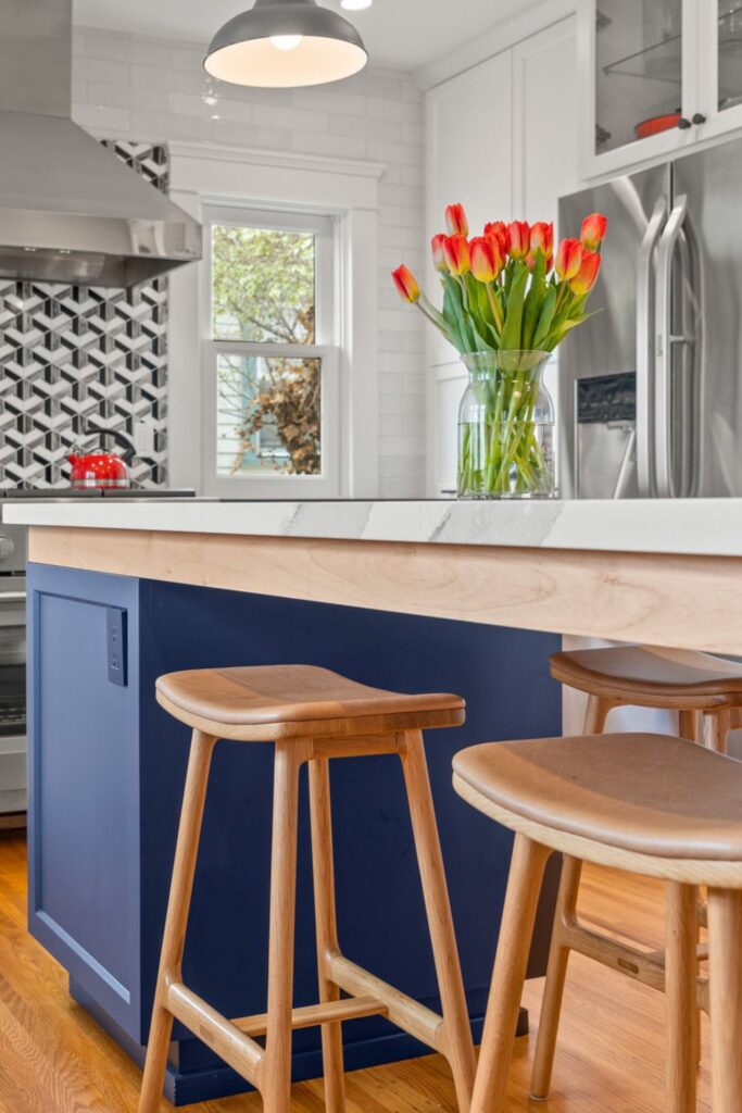 Blue kitchen island with wood trim, white countertop and wood stools beside stainless appliances and white cabinetry.