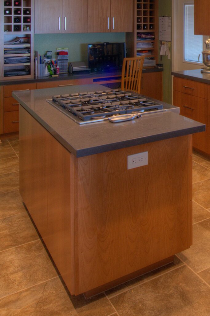 Kitchen cooktop island with gray countertop, wood base cabinetry and built-in desk with wine storage beyond