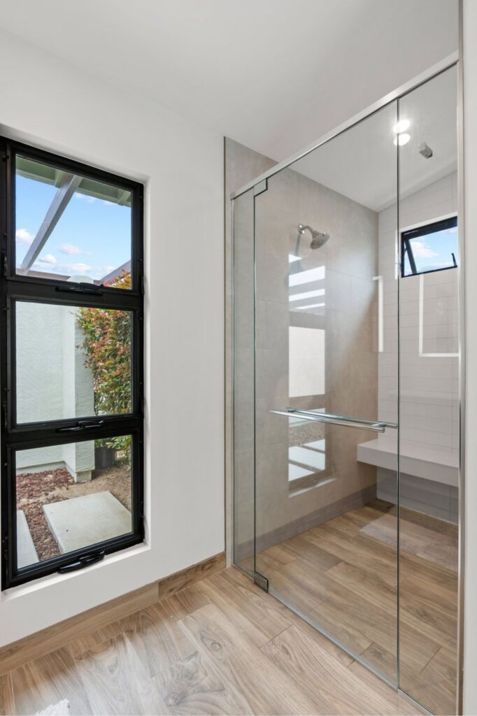 Glass-enclosed shower with gray wall tile, black-framed windows and wood-look flooring.
