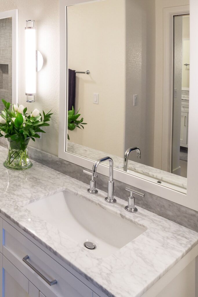 Primary bathroom vanity with white cabinetry, veined stone countertop, chrome faucet and large framed mirror.