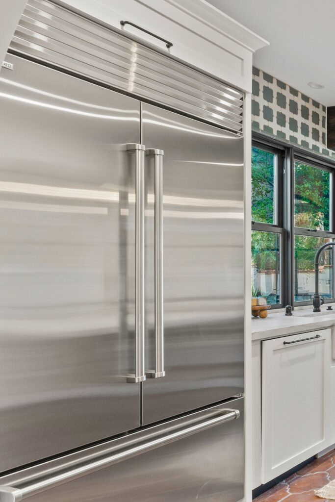 Stainless built-in refrigerator beside white cabinetry, black-framed windows, patterned wall tile and farmhouse sink