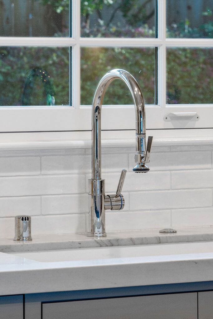 Chrome pull-down kitchen faucet beneath a divided-light window with glossy white tile backsplash.