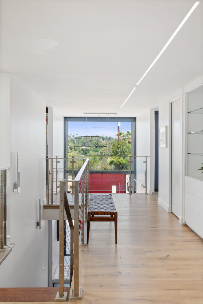 Upper hallway with wood flooring, cable railing, linear ceiling lights and a large window overlooking the entry.