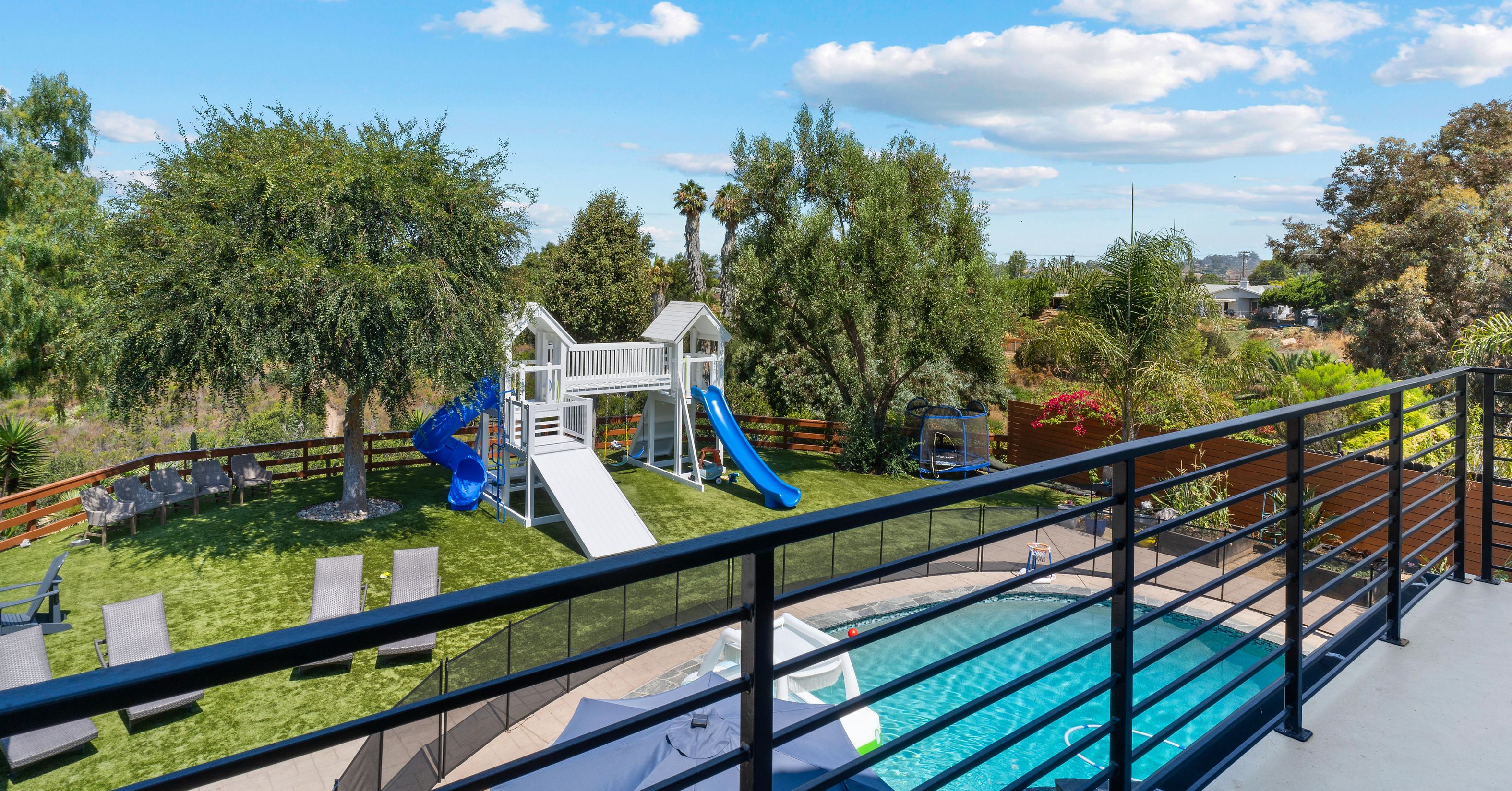 Upper balcony with black metal railing overlooking the backyard pool, lawn, play structure, and landscaping.