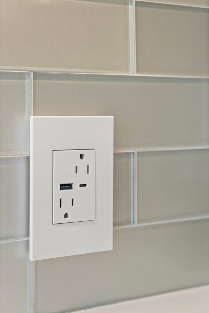 Kitchen electrical outlet with USB charging ports set into a light rectangular tile backsplash.