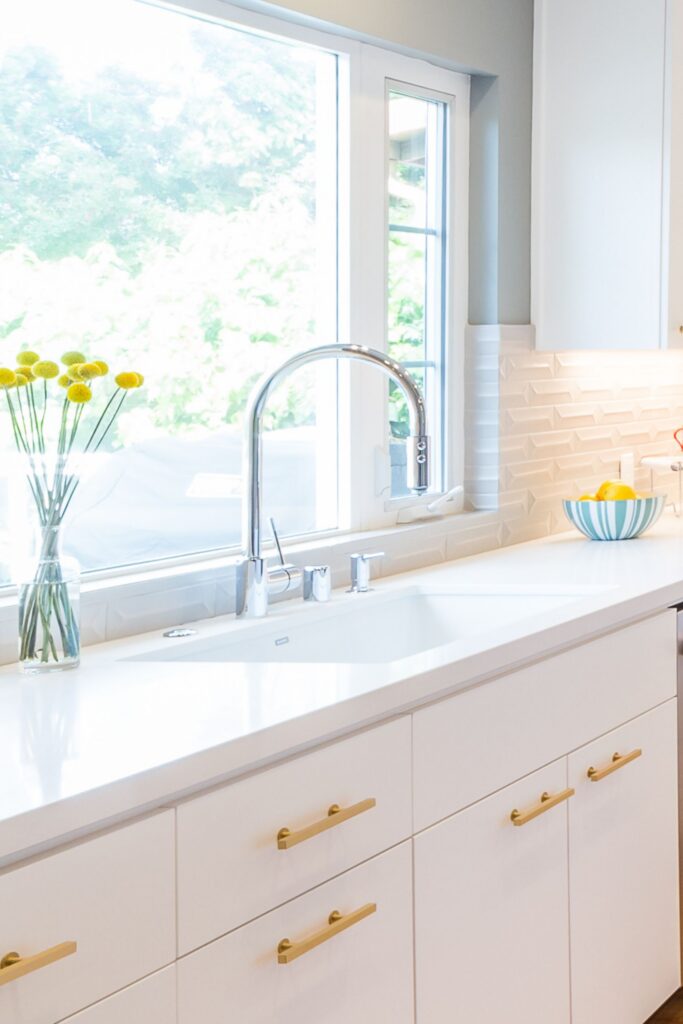 Kitchen sink beneath large window with white cabinetry, brass pulls and dimensional white tile backsplash