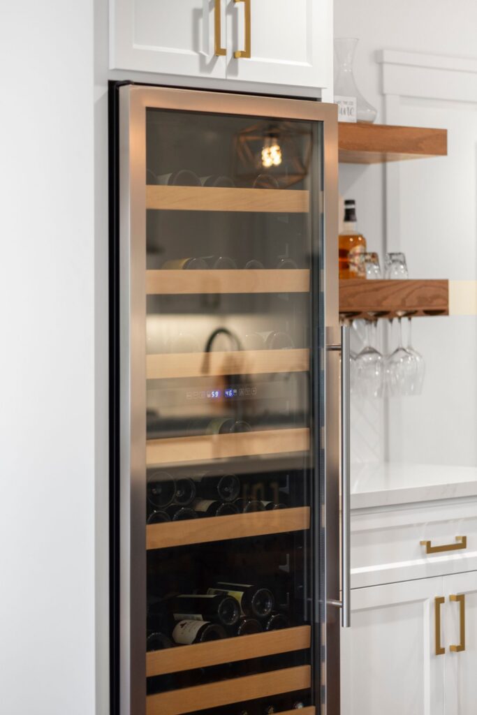 Tall glass-front wine refrigerator with wood shelves beside white cabinetry, floating shelves, and hanging stemware.