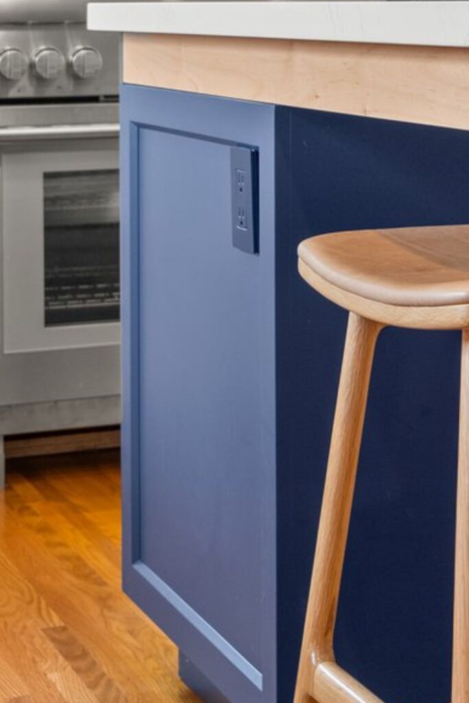 Close-up of blue island end panel with built-in outlet, wood stool, white countertop and hardwood flooring.