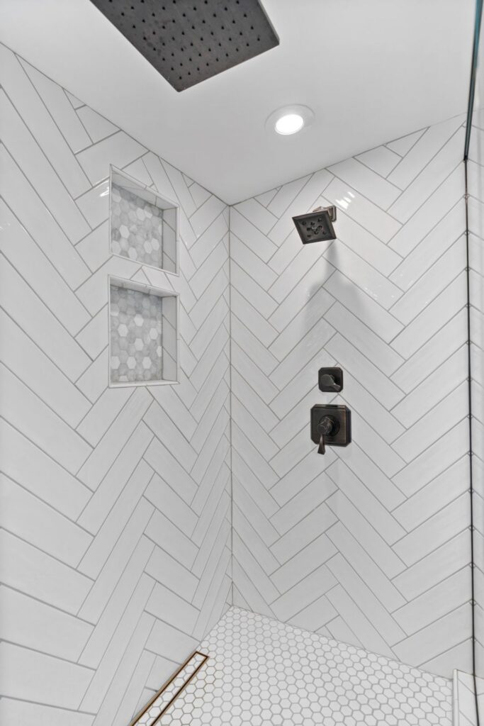 Walk-in shower with white herringbone tile, dark metal fixtures, recessed niches and hex mosaic shower floor
