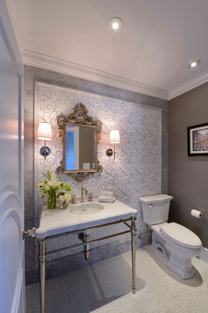 Powder room with marble console vanity, ornate mirror, wall sconces, toilet and decorative mosaic tile wall