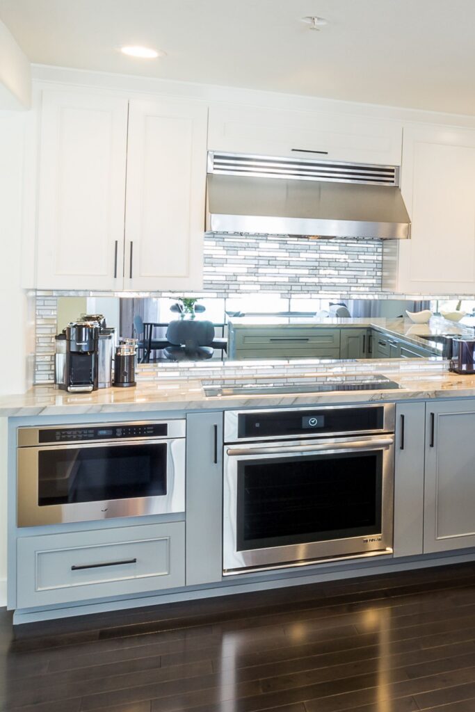 Kitchen appliance wall with white and gray cabinetry, stainless ovens, mirrored backsplash and mosaic tile accent.
