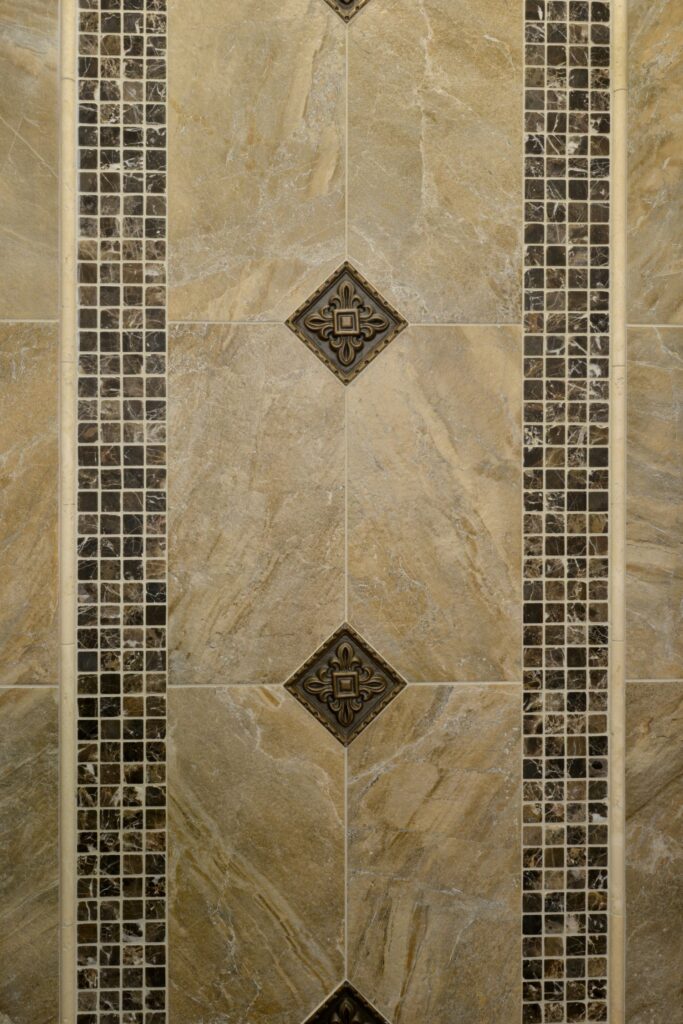 Bathroom wall detail with beige stone tile, dark mosaic bands and decorative metal accent tiles.