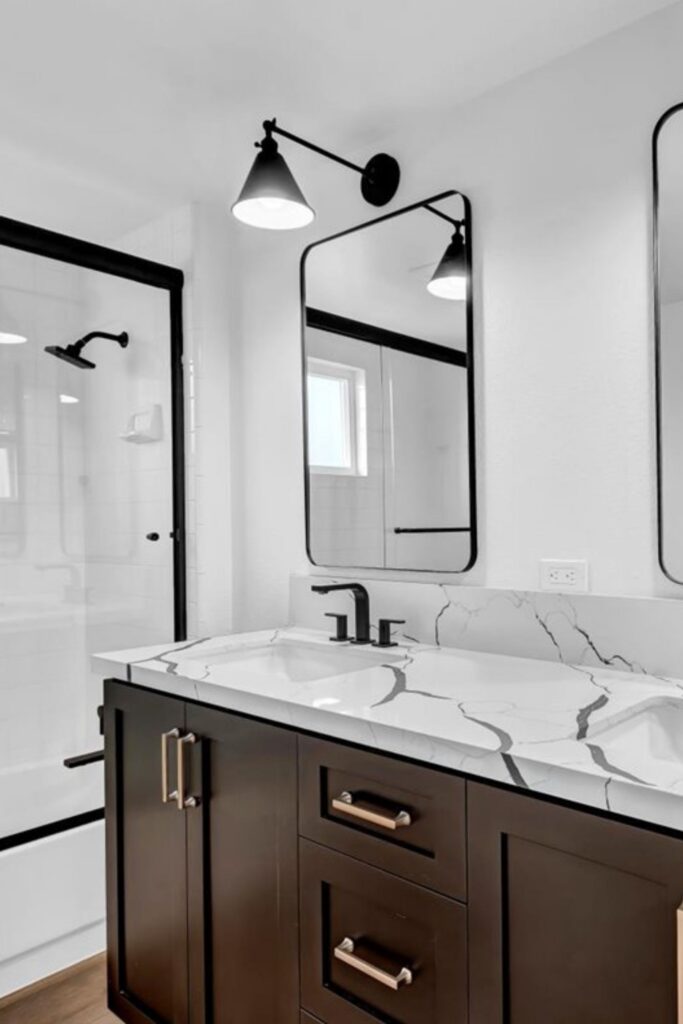 Bathroom vanity with dark wood cabinetry, veined white countertop, black fixtures, framed mirror and glass shower enclosure.