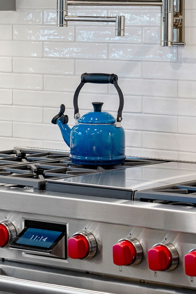 Stainless range with red control knobs, blue kettle, white tile backsplash and wall-mounted pot filler.